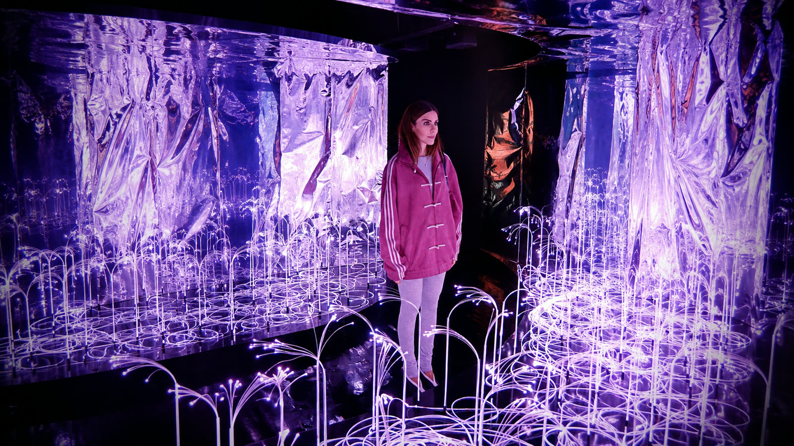 Samara Checon dentro da instalação de espelhos e luzes da exposição, vestindo casaco rosa